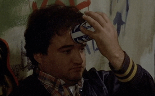 Movie gif. John Belushi as Bluto in National Lampoon’s Animal House leans against a graffitied wall and crushes a can of beer on his forehead.