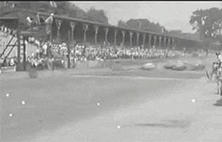 Video gif. Archival black and white footage of the Indy 500 shows vintage race cars speeding down a track as they kick up dirt.