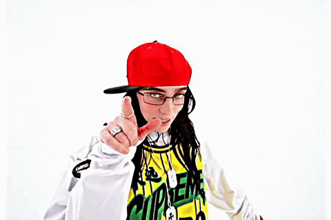 Music Video gif. From Billie Eilish's music video for "LUNCH", Billie is on a stark white background dressed in a white long sleeve shirt and a yellow and black jersey over the top, with a red flat brim hat on. She is pointing at the camera while singing her song and getting closer to the lens.