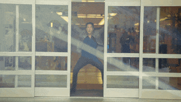 Music video gif. We see Pat Monahan from Train in Play That Song come through the automatic glass doors of a store. He dances out of the building towards us, pumping his pointing fingers in the air and then jumping up victoriously.