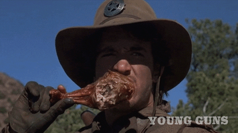 Movie gif. Dermot Mulroney as Dirty Steve Stevens in Young Guns. He's squinting at something far in the distance and as he gnaws on a big turkey leg.