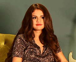 Celebrity gif. Selena Gomez raises her eyebrows and shrugs indifferently, sitting on a couch.