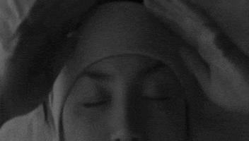 Video gif. Woman lays down in bed with her eyes closed. She pulls down a sleeping mask that has text that reads, “Let me sleep.” Over her eyes are drawings of closed eyes.