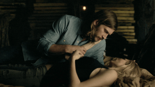 TV gif. Lying on a bed, Greyston Holt as Clay in Bitten holds the hand of Laura Vandervoort as Elena and kisses her on the mouth.
