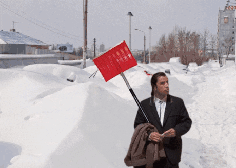 Movie gif. John Travolta as Vincent from Pulp Fiction superimposed on a snowy scene holds a brown coat and a red snow shovel. He looks around at the quiet suburban street, where several cars have been buried under snow, then takes another look around with a clueless shrug. 