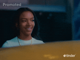 Sponsored gif.  Pink bubble letter text that reads "Blushing" flies into frame as a woman looks away from it with a smile, a shy or bashful expression on her face. The Tinder logo appears in the bottom right corner.