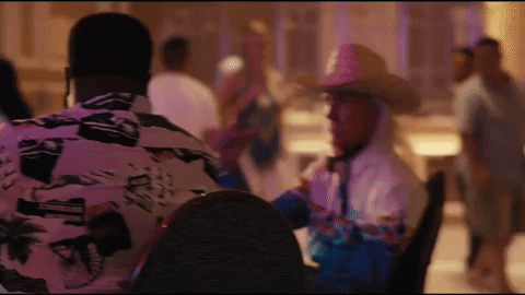 Music Video gif. Man with long gray hair and mustache wearing a cowboy hat sits at a gambling table in a casino and tips his hat at us in an Imagine Dragons music video.