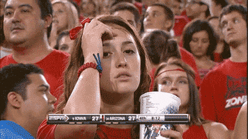 Sports gif. Young woman spaces out in the stands at an Iowa vs. Arizona college football game until she notices the camera on her and awkwardly snaps out of her daze by smiling and cheering loudly.