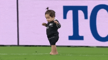 Video gif. Toddler with a tiny pony tail wanders barefoot across a rugby field.