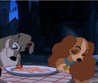 Disney gif. A clip from the iconic spaghetti scene from Lady and the Tramp. Using his nose, Tramp nudges a meatball across a dinner plate towards a flattered Lady.