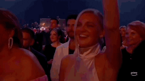 ACM Awards gif. Woman in pink halter mock neck bears a happy expression with one arm raised as she vibes to the live performance.