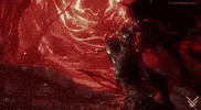 Video game gif. An intense, dark fantasy scene with a human-like figure with dragon wings and textured armor standing amidst swirling red fire and embers. The background features chaotic, fiery light patterns and twisted, shadowy shapes. The dark figure quickly outstretches its hand as if casting a spell.