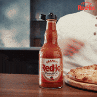 Sponsored gif. Smiling man shakes Frank’s RedHot sauce onto a pizza. A red chef’s scarf suddenly lands around his neck, a brick column appears to the right of him, and a red and white checkered table cloth and lit candle slide onto the table in front of him. Text reads, “Ciao!”