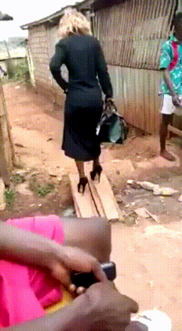 Video gif. Woman in all black and tall heels walks carefully across wood planks and down a dirt path before trying to speed up and stumbling, her ankles twisting back and forth with each step as she tries to regain her balance.