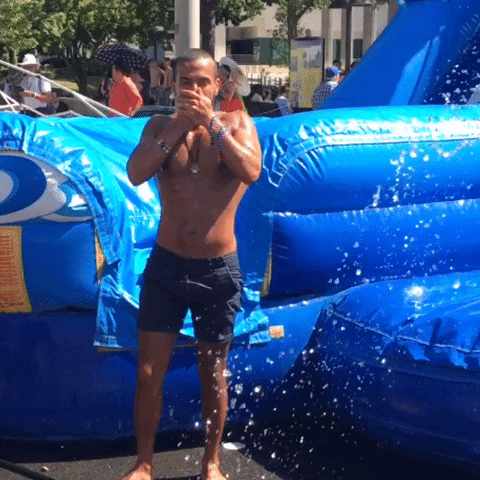 Video gif. Man has just come off an inflatable wet slide during the pride parade and he gets sprayed with a hose.