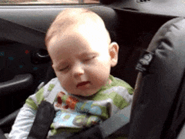 Video gif. Baby sleeps in a car seat then suddenly sits up with a gleeful smile.