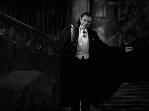 Movie gif. Bela Lugosi as Count Dracula in the 1931 Dracula stands in front of a grand, haunting staircase and spreads his cloaked arms out, saying, "I bid you welcome," which appears as text. The only source of light comes from the candle he holds, and a creepy smile plays upon his face.