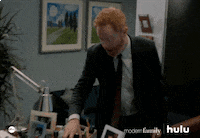 TV gif. Jesse Tyler Ferguson as Mitchell on Modern Family. He picks up his coffee mug and tries to leave but gets called back so he does a smooth 360 turn and leans his head in to listen.