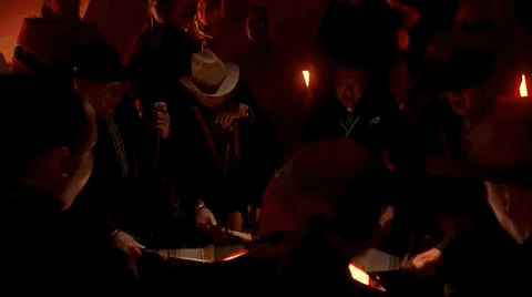 Oscars 2024 gif. Scott George and Osage singers perform 'Wahzhazhe (A Song For My People)' from Killers of the Flower Moon. Men in cowboy hats are obscured in the shadows while they circle around and beat on a powow drum bathed in the spotlight. 