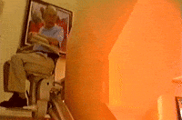 Video gif. A man is riding on a personal stair lift in his home but the stair lift slowly descends him into a fiery pit.