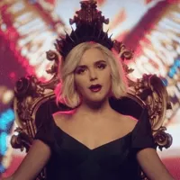 TV gif. Kiernan Shipka as Sabrina Spellman in the Chilling Adventures of Sabrina, sitting on a throne and winking.