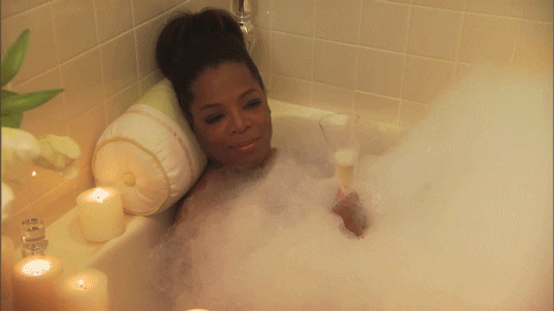A GIF of Oprah relaxing in a bubble bath. She's drinking champagne and smiling, and she's surrounded by candles.