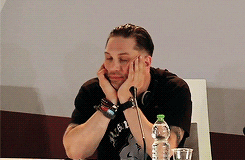 Celebrity gif. Tom Hardy sits behind a microphone in a press conference leaning his chin into his hands, looking bored. The text, "I'm bored," continuously flashes at the bottom.