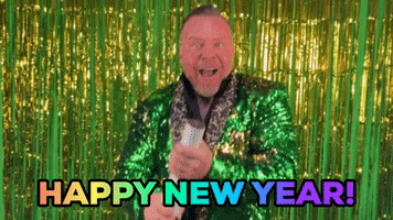 Video gif. Man wearing a sparkly green suit in front of a golden and green shiny background. He smiles at us and holds a confetti in his arms, letting it pop. Confetti falls in front of him. Text, “Happy New Year!”
