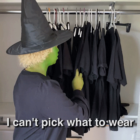 Video gif. Woman painted green and wearing a witch hat and a black dress looks around from a closet full of black identical clothes, annoyed. She slaps her side and says, “I can’t pick what to wear.”