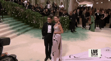Met Gala 2024 gif. Closeup of Lily James posing next to her date, quickly striking different poses for the camera. She's wearing a pale pink Erdem gown with a fitted bodice with a scalloped plunging neckline. A black vine with leaves detail weaves up and over her shoulders and her hair is swept back in a mid bun.