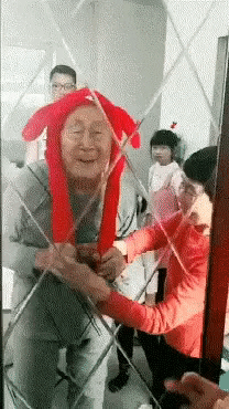 Video gif. Old man is delighted to discover that the ears on his red hat pop up when he pulls the strings on the side. He giggles, and his family reacts in laughter.