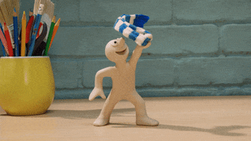 Stop motion gif. Chas from The Epic Adventures of Morph stands on a desk with yellow cup of school supplies and happily twirls his blue and white scarf in the air.