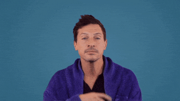 Celebrity gif. Wearing a blue zip up sweater against a lighter blue background, a pensive Simon Rex holds a hand to his chin, and nods as if he's mulling over some info.