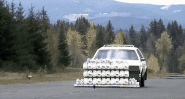 Video gif. Large white car with a flat front plows through a stack of white cartons, releasing a humongous explosion of white into the air as the cartons fall upon the road.