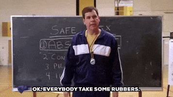 Movie gif. Standing in front of a chalkboard that says "Safe Sex" with the word "abstinence" underlined, Dwayne Hill as Coach Carr on Mean Girls bends down to pick up a box of condoms. Text, "Ok, everybody take some rubbers."