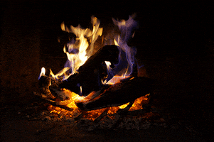 Video gif. Dark, moody fireplace lit by a roaring fire.