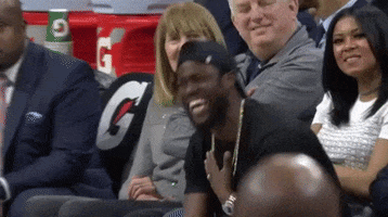 Celebrity gif. Kevin Hart sits on the sidelines at a basketball game holding a hand to his throat as he laughs uncontrollably. 