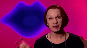 Reality TV gif. Contestant on RuPaul's Drag Race viciously hisses at us and clenches their hands into claws. They sit against a hot pink backdrop decorated with a dark blue lip.