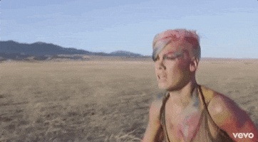 Music video gif. From the video for Try by Pink, the singer Pink runs in a flat grassy field, wearing a green bra, skin dappled with colors.