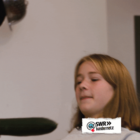 Video gif. In a kitchen, a smiling girl holds out a cucumber like a limbo stick while another girl skillfully bends backward and limbos beneath it.