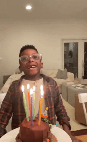 Video gif. Young boy with clear framed glasses opens his mouth wide and takes a big breath as he blows out the candles on his birthday cake.