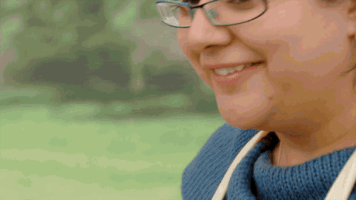 Reality TV gif. A contestant on the Great British Bake Off shyly smiles and throws us a pair of crossed fingers and the emoji crossed fingers pops up next to her as well.