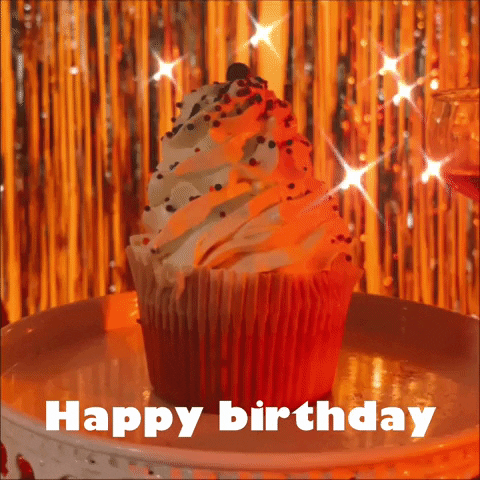 Video gif. A cupcake with frosting piles high sits on a plate in a dimly lit space with metallic streamers twinkling in the background. Text, "Happy birthday."