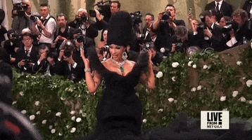 Met Gala 2024 gif. Slow motion closeup of the top of Cardi B's black strapless Windowsin gown. She brings her long pointy emerald green nails slowly down to her sides. The black wing panels emerging from her waist flare out in a v-shaped pattern that ends just above her shoulders and forming the wide v-neckline of her dress.